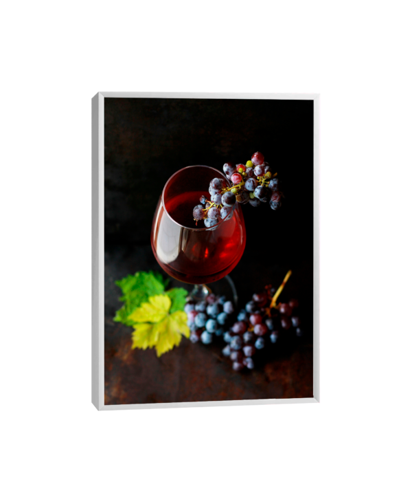 Quadro que retrata uma taça de vinho tinto em cristal sobre uma mesa de madeira rústica, acompanhada por cachos de uvas roxas e folhas verdes, evocando sofisticação e charme.